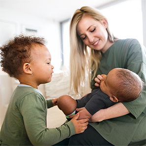 A mom is holding her baby in her arms, smiling. She is feeding the child a bottle. A slightly older child is standing next to them looking curiously at the baby.