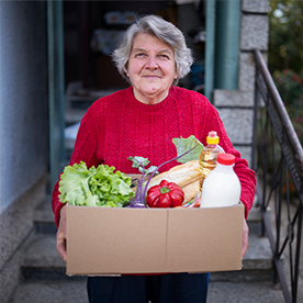 An elderly white woman stands outside, looking hopeful at the camera, holding a box of fresh fruits and vegetables.