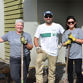 Two women and one man are standing outside, smiling at the camera. The two women are holding shovels in their hands, all three individuals are wearing gloves.