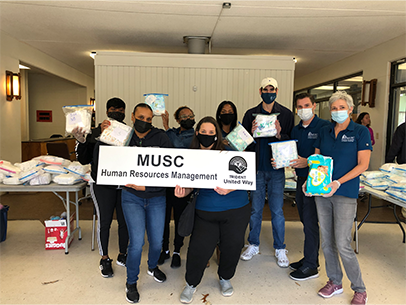 Several volunteers wearing MUSC shirts pose for a photo at the Bundles of Joy Diaper Bank. They are holding a plastic ziploc bag filled with diapers, and are holding a sign that says MUSC Human Resources Management.