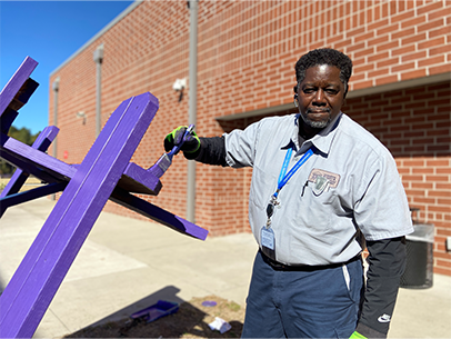 A volunteer is at a school painting a picnic table bright purple, volunteer is looking at the camera smiling