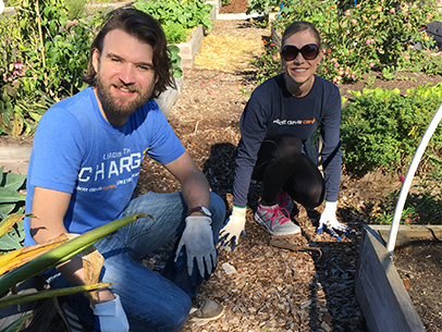 Two volunteers wearing Elliott Davis volunteer shirts crouched on the ground to garden, both looking at camera and smiling