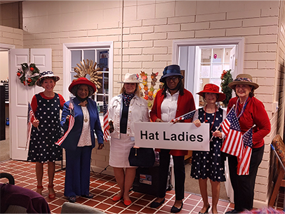 Six women at the Charleston Senior Center wearing outfits of red, white and blue and fun, festive hats. They are posing for a photo, smiling, several hold small USA flags, and one is holding a sign that says Hat Ladies
