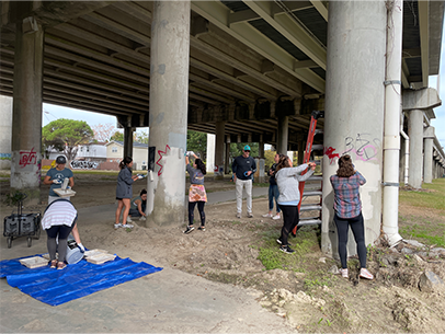A large group of volunteers are painting over grafitti on poles under an overpass in downtown charleston