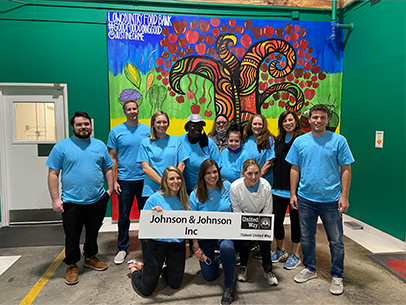 A large group of volunteers wearing bright blue Days of Caring shirts pose in front of a colorful mural at Lowcountry Food Bank. Three individuals are kneeling in the front for the photo, holding a sign that says Johnson & Johnson Inc
