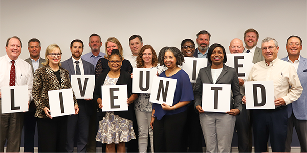 A photo of several volunteers wearing professional attire. Individuals hold a sign in their hands with a letter on it. The letters all spell out LIVE UNITED