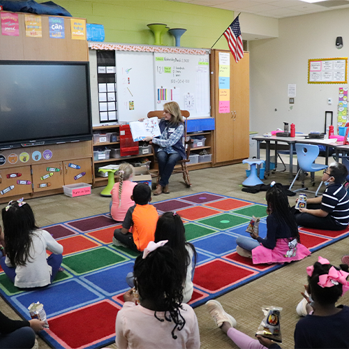 Photo of volunteer reading to a group of students in a classroom. The students are sitting on the floor.