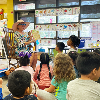 An older woman sits in a rocking chair, she has on a straw beach hat, blue glasses and floral dress. She is holding up a book cover and showing it to a classroom full of students between the ages of 6-8.