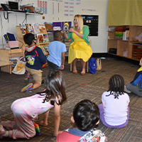 A volunteer wearing a bright yellow dress and green cardigan reads a book to a classroom. She is holding the book for the students to see the illustrations, and one student is looking closely at the illustrations