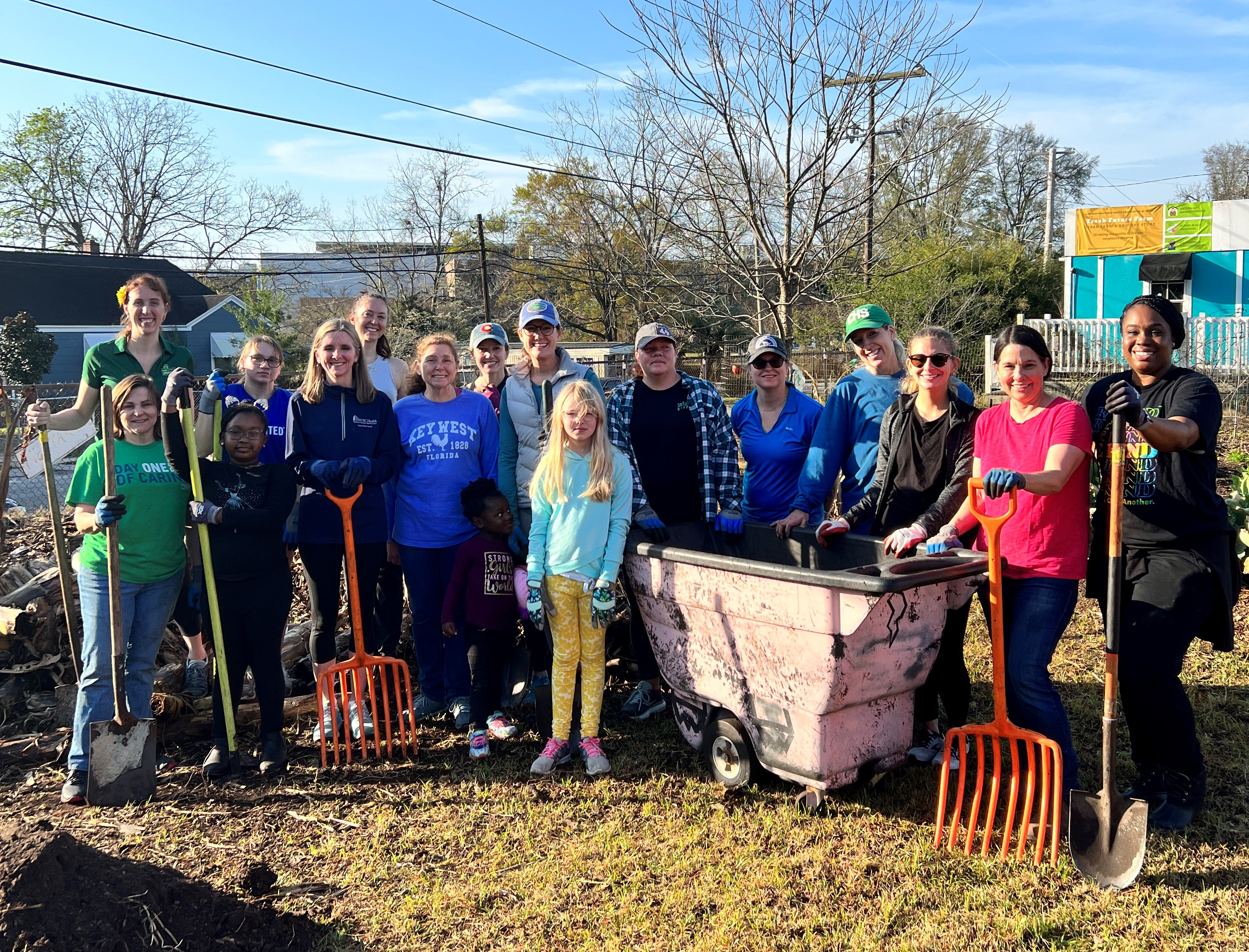 "A large group of volunteers stands outside on a field and smiles, many holding rakes or shovels."