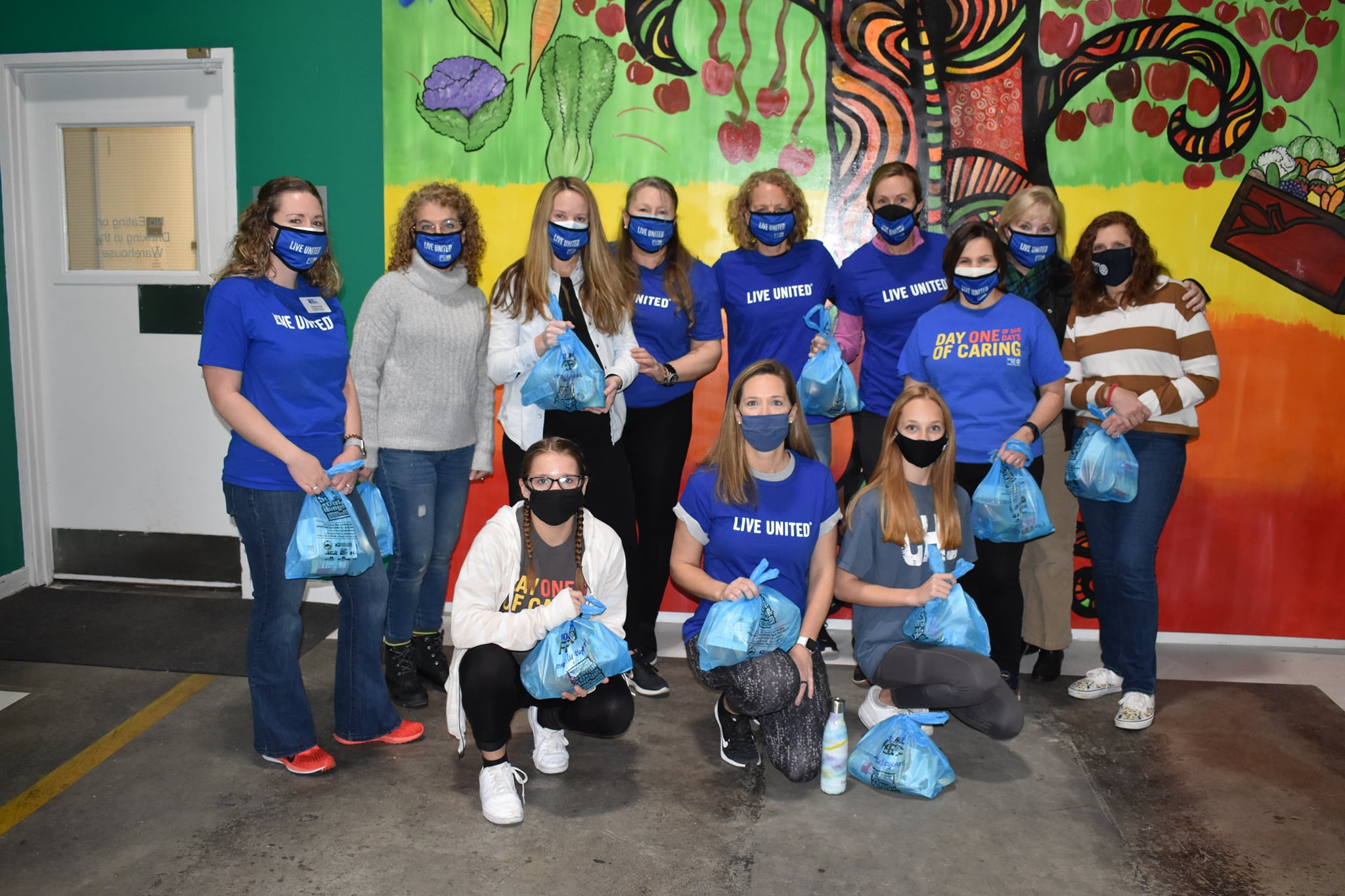 “A group of volunteers wearing Live United masks stands holding blue bags in front of a mural, which features a painted tree and fresh produce.”