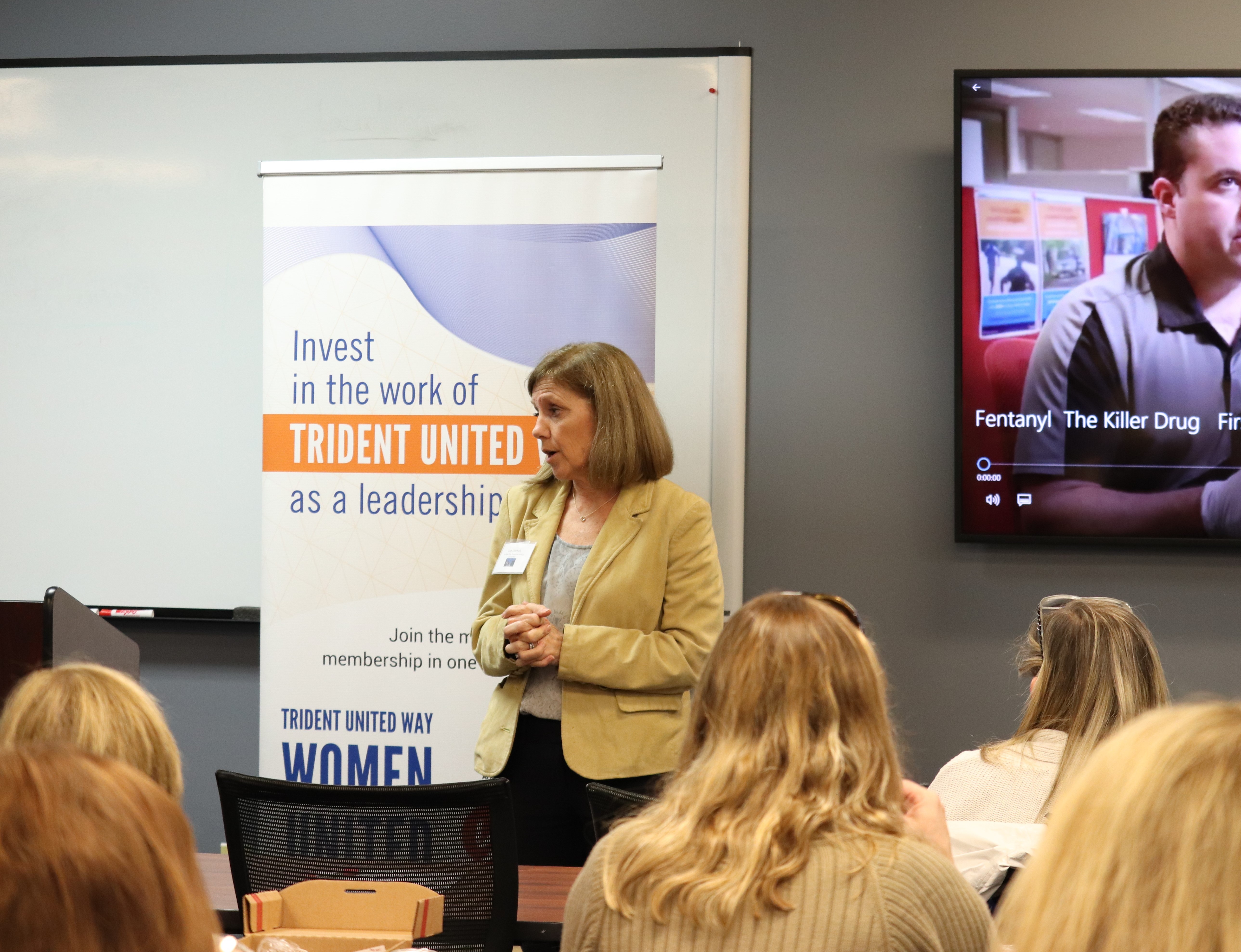 “Speaker Lisa Mitchell stands in front of a group of women, in front of a sign that reads ‘Invest in the work of Trident United as a leadership.’ A video plays on the TV to her right, with the description ‘Fentanyl, The Killer Drug.’”