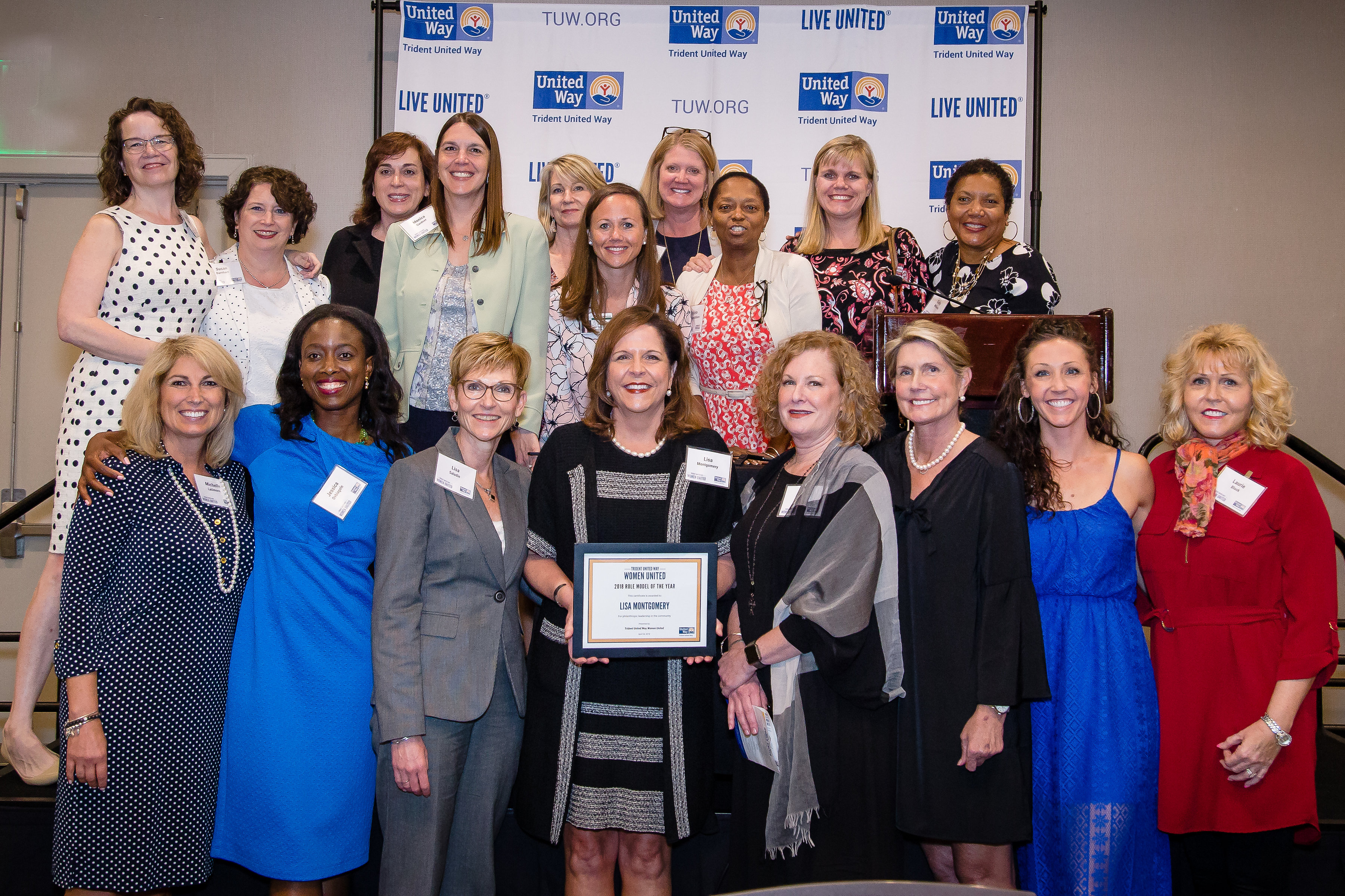 "A group of women stands in front of a Trident United Way background. The woman in the middle holds up an award certificate which reads '2018 Role Model of the Year: Lisa Montgomery.'"