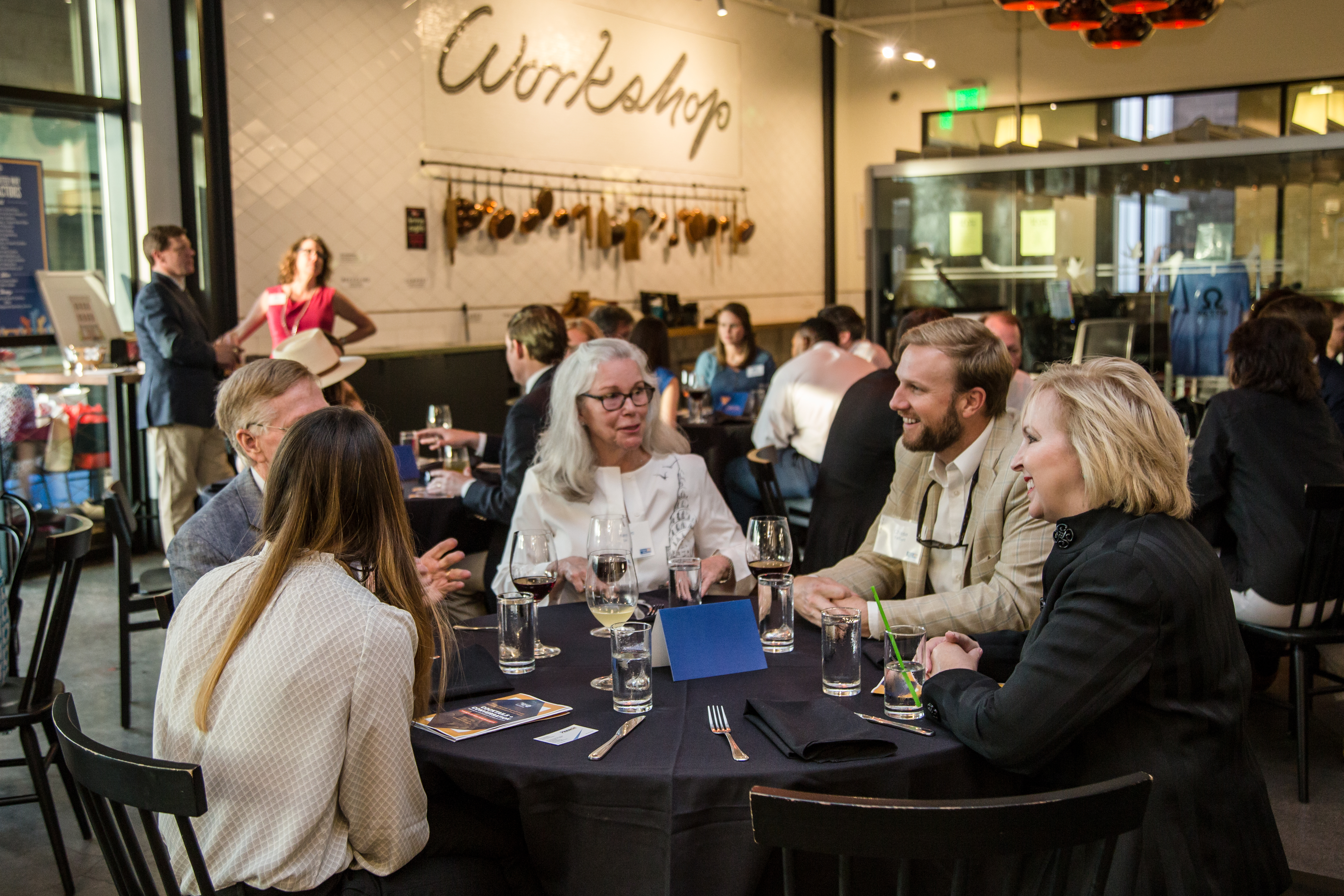 "A group of attendees sits around a round table and discusses, with similar tables seen in the background. The wall features the word 'Workshop,' underneath which hang a number of cooking supplies." 