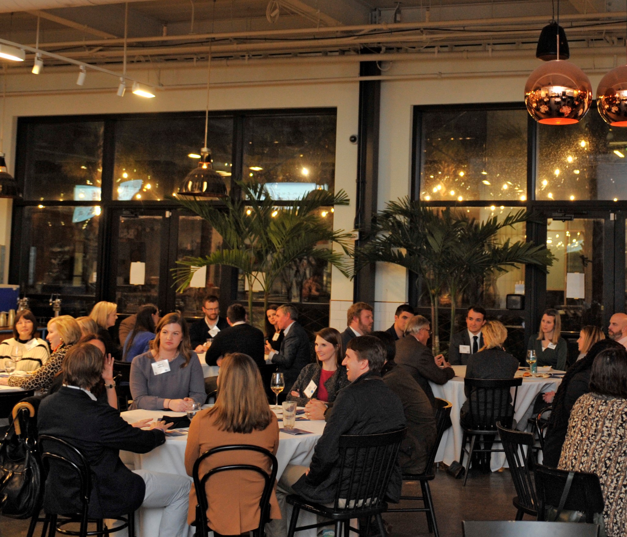 "A large group of people sits at multiple tables inside of a nice building with large windows revealing the dark outside."