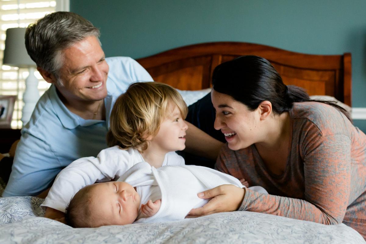 Photo of a family of four laying on a bed, a man and woman are looking at a younger boy who is holding a baby