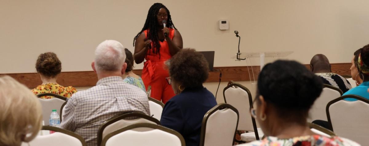 A woman stands in front of a room, speaking to a group of diverse people 