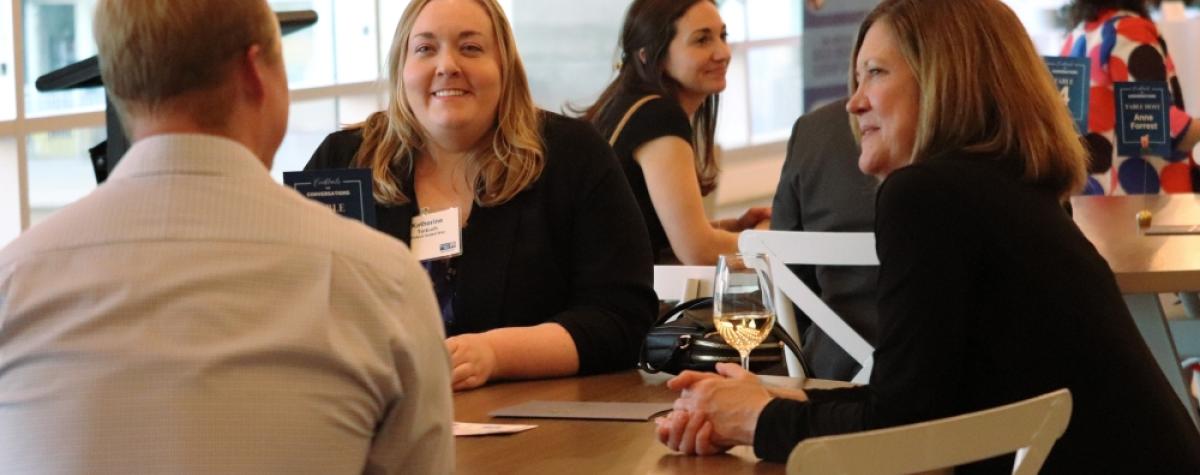 Two women are sitting at a table, both are smiling and looking at the man sitting at the table with them. There are groups of tables behind them in the room