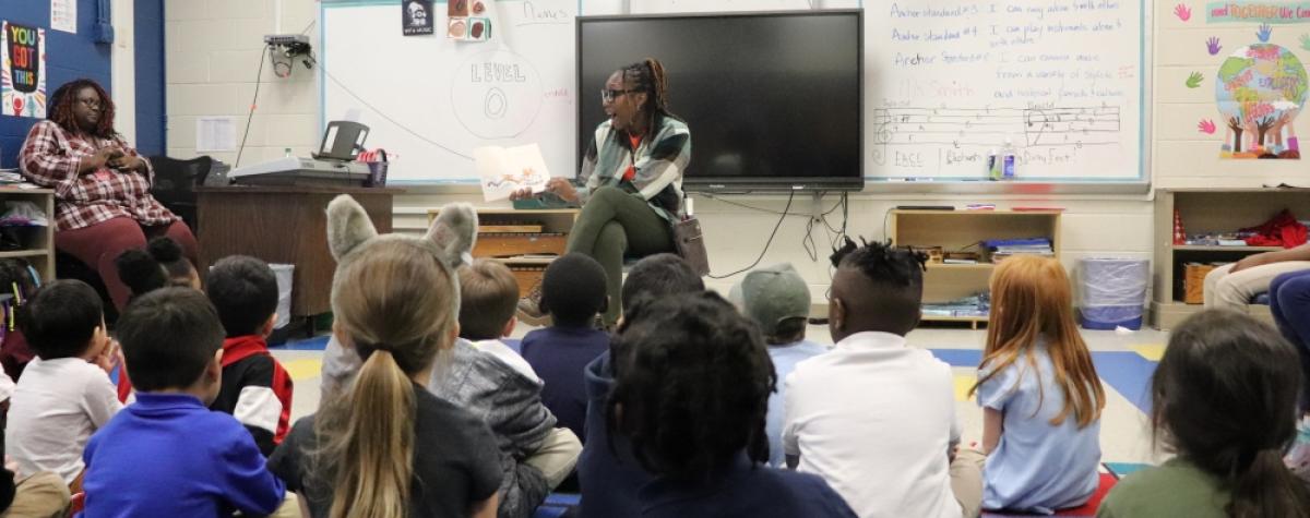 A woman sits on a chair in a classroom, all the children are sitting in front of her on the floor. She is in the middle of reading a book aloud to them with excitement