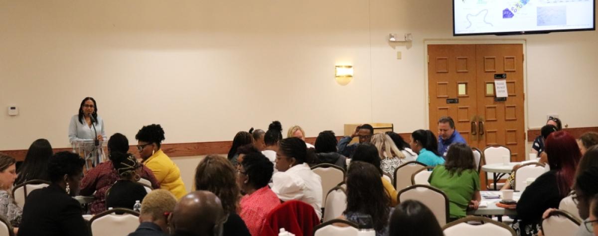A full room with people sitting around tables, a person stands at the head of the room speaking into a podium mic