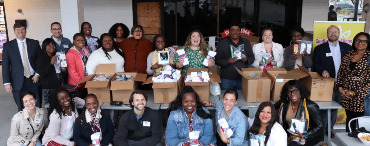 A group of people from a past sip and serve are posing for the camera standing near boxes of packed supplies and smiling