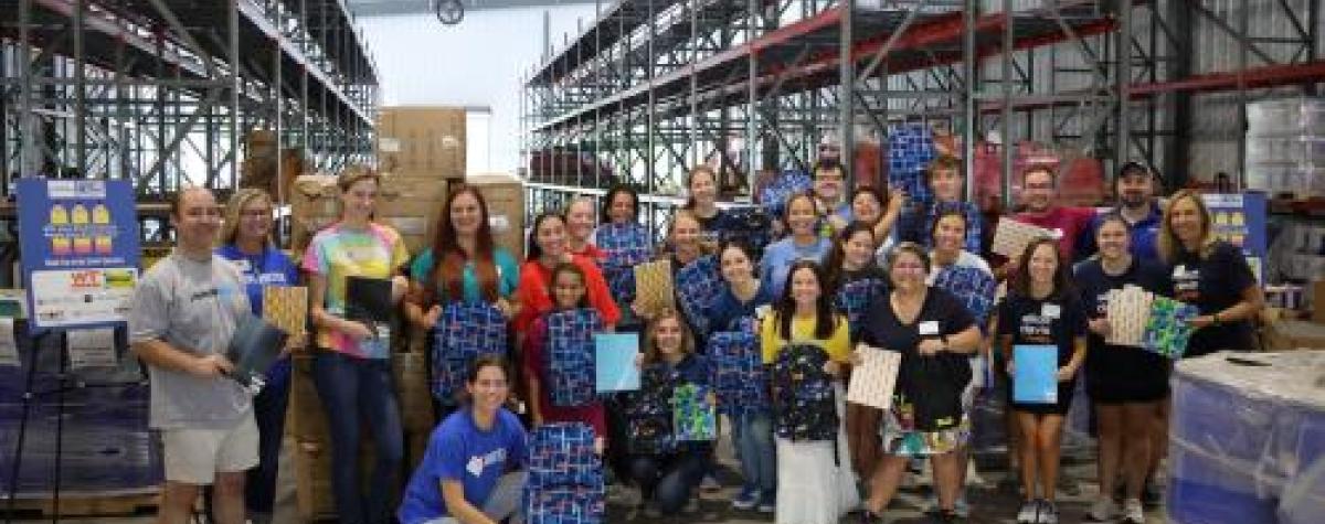 A group of 30 volunteers pose in a warehouse setting holding backpacks and school supplies