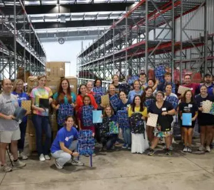 A group of 30 volunteers pose in a warehouse setting holding backpacks and school supplies