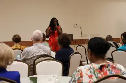 A woman stands in front of a room, speaking to a group of diverse people