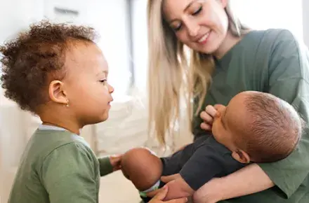 A woman is holding a baby in her arms, she is smiling looking down at him. A younger child stands next to the baby looking over it.