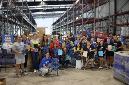 A group of 30 volunteers pose in a warehouse setting holding backpacks and school supplies