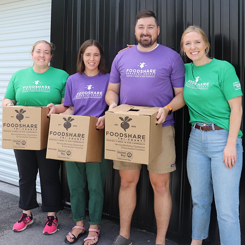 Four individuals standing outside wearing FoodShare Berkeley County t-shirts. Two are wearing green shirts, two are wearing purple shirts. Three are holding boxes that say FoodShare Tri-County on the outside.