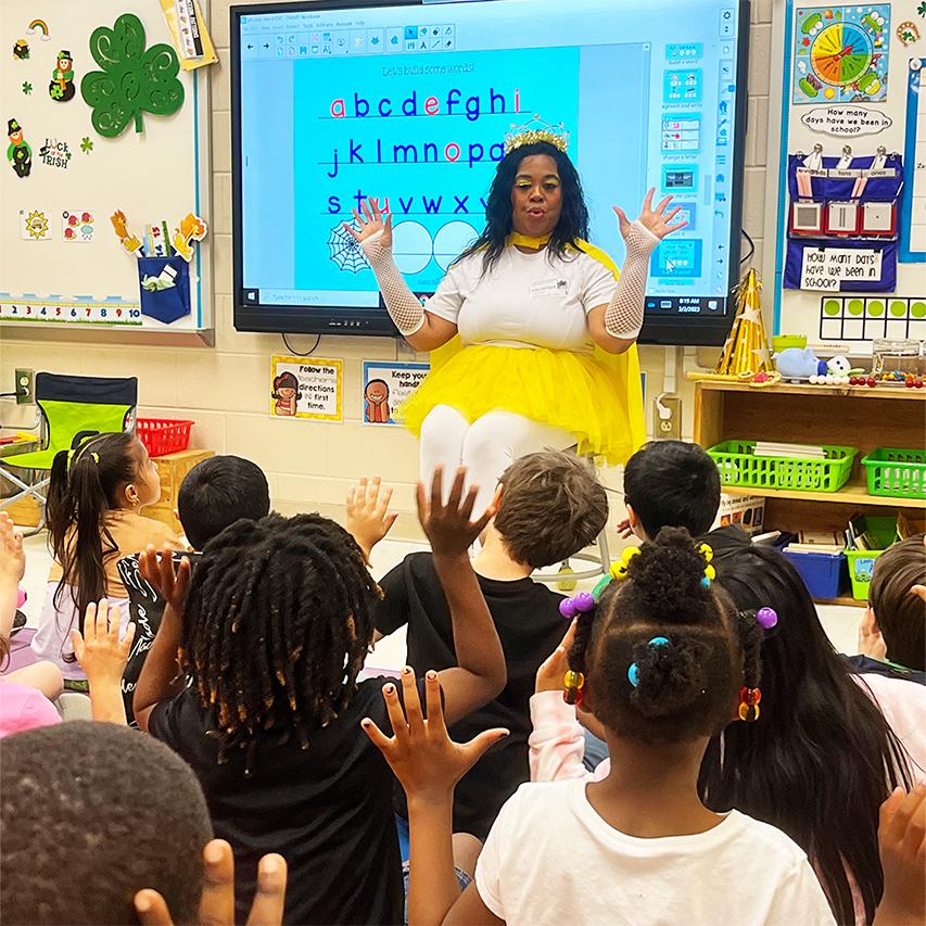 Woman wearing a yellow tutu, tiara, holding her hands up, facing a classroom. Children in classroom facing woman holding their hands up to mimic her.