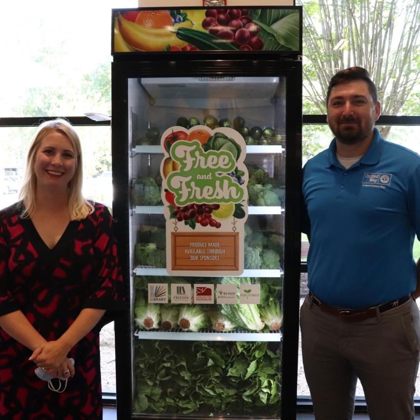 Photo of two individuals standing next to a clear fridge filled with vegetables that says "Free and Fresh" on it