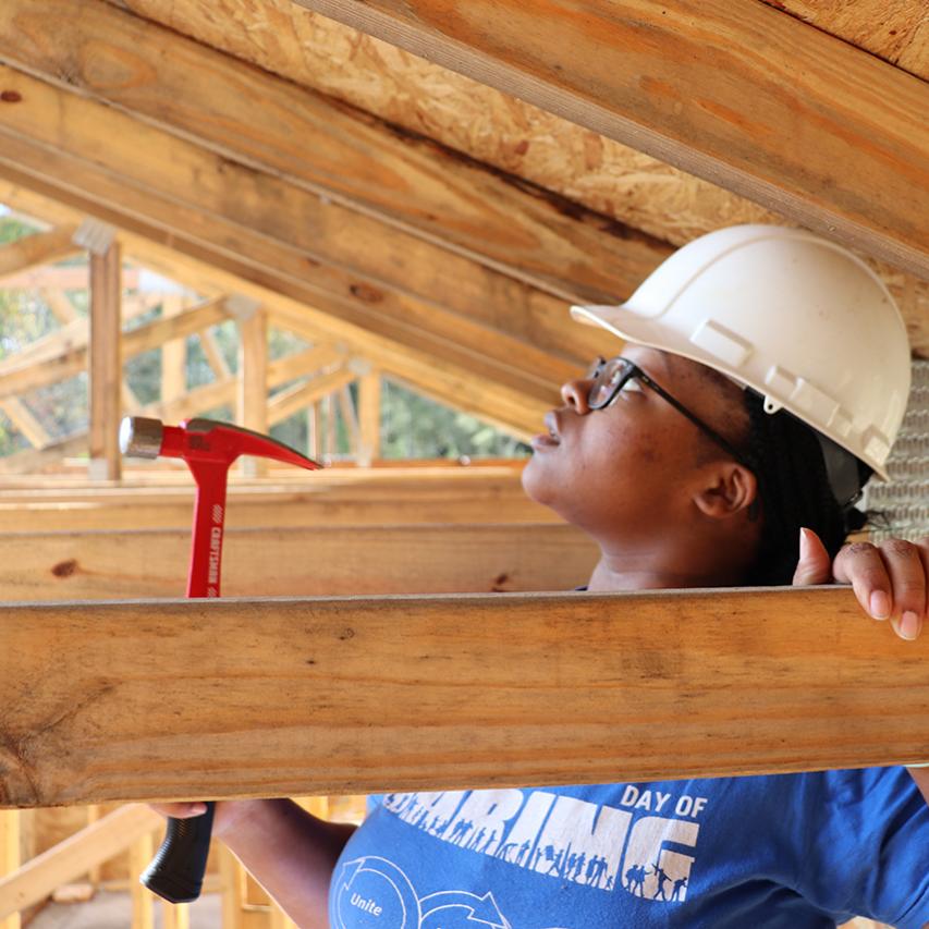 Photo of a woman looking up at wooden beams wearing a hard hat and holding a hammer