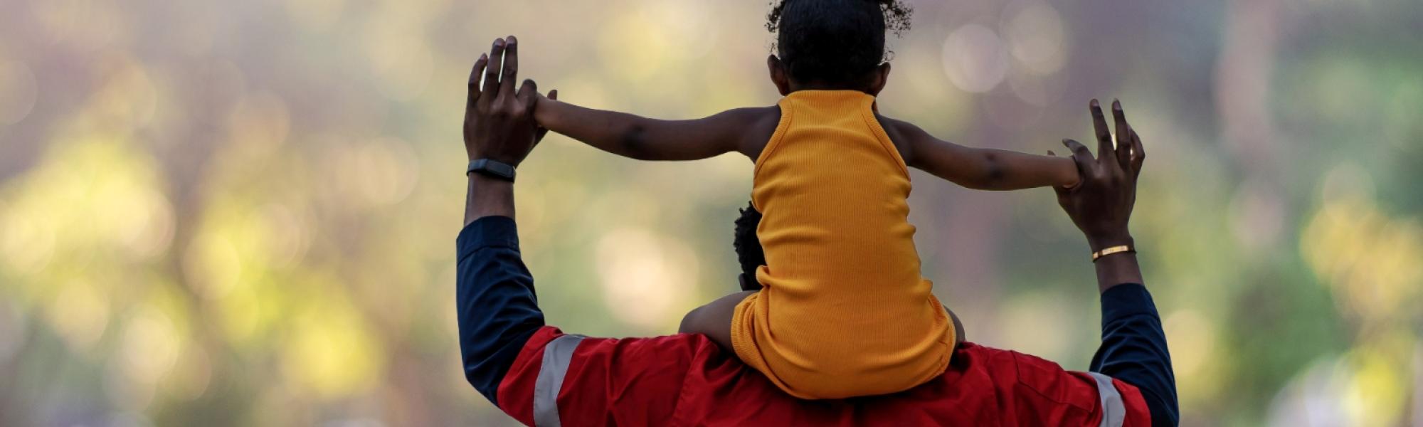 Adult with child on shoulders outside with their backs to us. They are holding hands in the air.