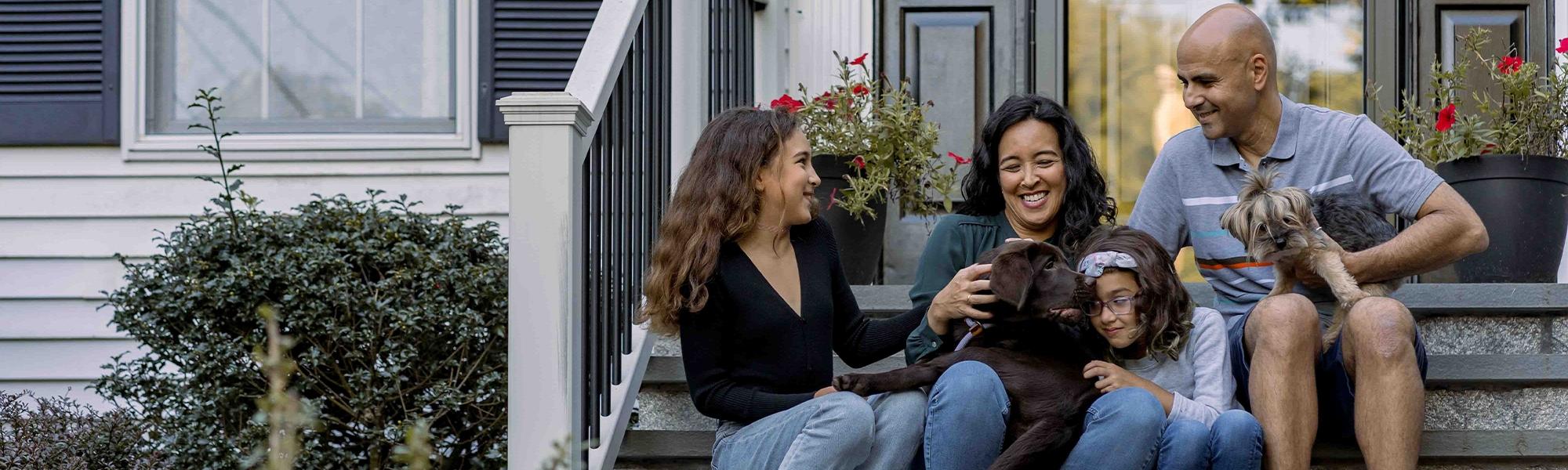 A family of four sits in front of the house playing with their two dogs