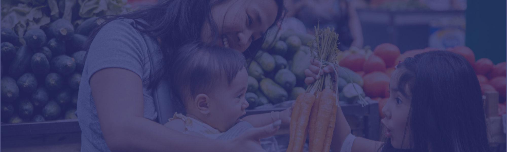 A woman is standing in the store with her 2 children, one son and one daughter. They are smiling and holding the produce while putting it into the cart together.