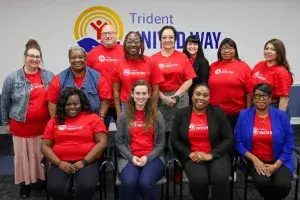 Trident United Way Family Coaches posing for a photo against a white wall with the TUW logo on it. They are all wearing red TUW shirts, some standing and some seated in chairs, all smiling at the camera.