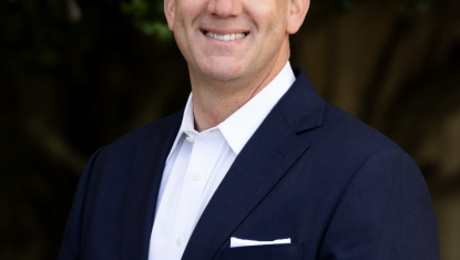 David stands in front of a tree smiling in a blue suit, white shirt underneath with no tie. He has a white pocket square.