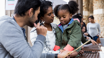 A family sits on a bench in a building holding a clipboard, a child sits on the mother's lap.