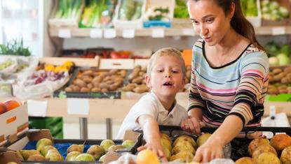 A mother and son are at a grocery store in the produce section. The son is reaching for a piece of fruit with determination on his face. The mom is helping him.