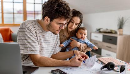 A man, woman and their child sit behind a laptop computer while in their home. They are looking at financial paperwork while the baby sits on the mother's lap.