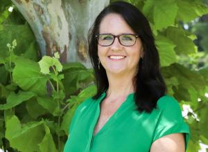 Photo of Amy Maciariello outside in front of a tree. She is wearing a kelly green v-neck blouse and black glasses. Amy has shoulder length straight dark hair.