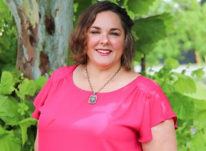 Photo of Christy Boudolf outside in front of a tree. She is wearing a dark pink blouse with a silver pendant necklace. Christy has short curly brown hair. 