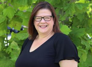 Photo of Helen Stungurys outside in front of a tree. She is wearing a black short sleeve blouse and black glasses. Helen has shoulder length straight dark hair.