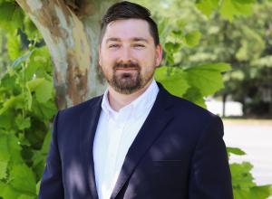 Photo of Joey Current outside in front of a tree. He is wearing a navy suit jacket with a white button down shirt. Joey has short dark brown hair and a beard. 