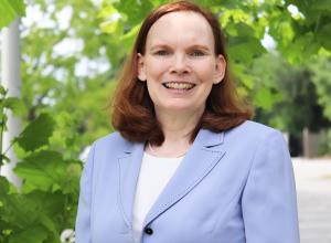 Photo of Maureen Feely outside in front of a tree. She is wearing a light blue blazer with a white undershirt. Maureen has reddish-brown short shoulder length hair.