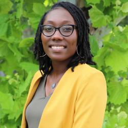 Photo of Angie Williams outside in front of a tree. She is wearing a olive shirt with a yellow cardigan and black glasses. Angie has short dark hair. 