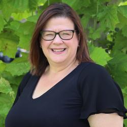 Photo of Helen Stungurys outside in front of a tree. She is wearing a black short sleeve blouse and black glasses. Helen has shoulder length straight dark hair.