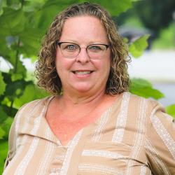 Photo of Kathe Faretra outside in front of a tree. She is wearing a tan and white striped blouse and thin glasses. Kathe has short curly brown hair. 